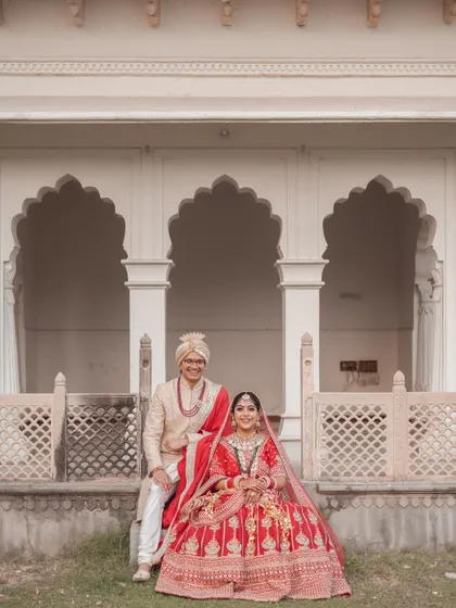 Seated against the elegant arches of the palace, the couple shares a quiet, happy moment. This portrait captures their regal attire and the serene atmosphere of their special day.