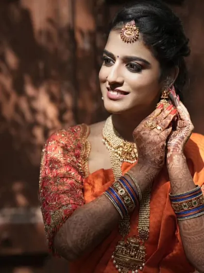 A candid shot of a bride adjusting her earring. She is wearing a beautiful temple jewellery set with her orange silk saree.