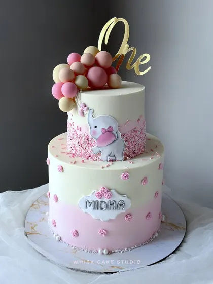 A lovely pink and white first birthday cake for a baby girl. This two-tiered cake is decorated with a cute elephant, balloons, and delicate sprinkles.