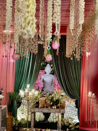 A serene and spiritual corner. We placed a Buddha statue behind a console table, framed by soft pink and green drapes and a ceiling of hanging flowers, creating a tranquil space.