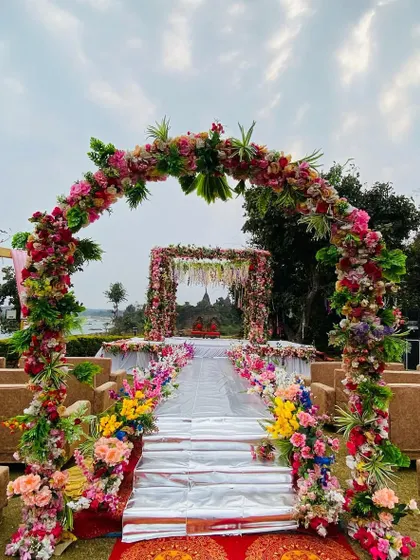 A breathtaking outdoor mandap with a full floral archway, set against a beautiful riverside view. The aisle is lined with colorful flowers.
