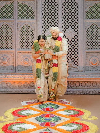 The couple stands on a beautiful floral rangoli. This view highlights the full length of their coordinated cream and gold wedding outfits.