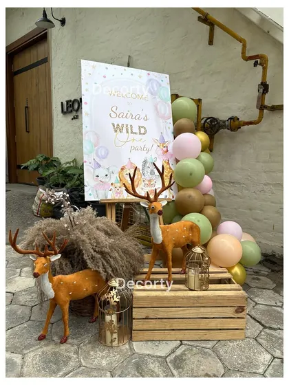 A charming welcome sign for 'Saira's Wild One Party'. The entrance is styled with rustic wooden crates, pampas grass, and adorable deer props to set a woodland safari tone.