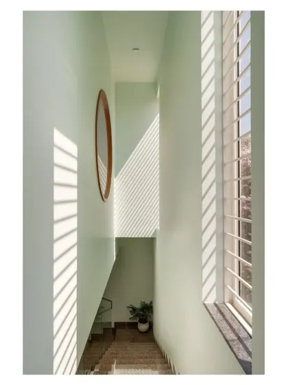 Sunlight streams through a large window, casting strong linear shadows down a narrow, double-height stairwell, creating a dramatic and ever-changing pattern on the walls.