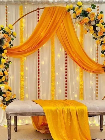 A modern and chic Haldi seating arrangement with a circular arch decorated with yellow drapes and floral bouquets. The white bench and fairy light backdrop add a touch of elegance to the traditional yellow theme.