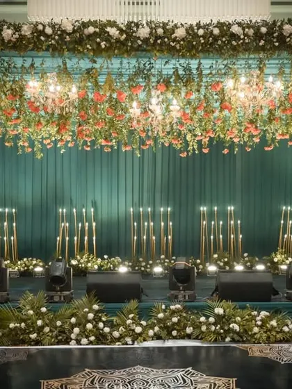 A luxurious stage design featuring a dramatic ceiling of hanging florals and chandeliers over a teal-paneled backdrop. The warm glow of candle-like floor lamps adds a layer of sophistication, ideal for a formal corporate dinner or awards ceremony.
