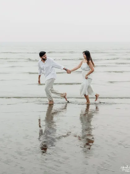 A classic romantic shot of a couple walking hand-in-hand in the shallow waters of a Mumbai beach. The reflection in the wet sand adds a beautiful, symmetrical element to the composition.