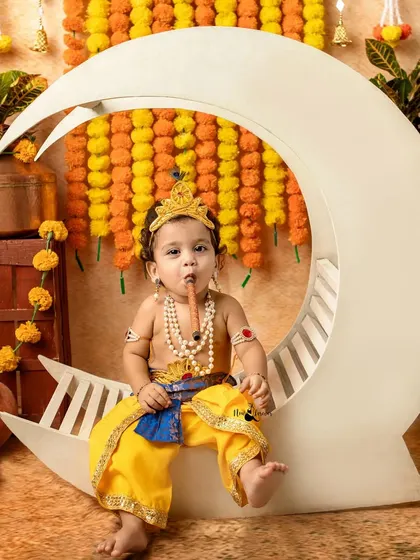 A fun shot of a baby as Krishna, playing his flute while sitting on the moon prop.