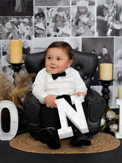 A classic first birthday portrait. This little gentleman is sitting in a mini leather armchair against a backdrop of family photos, symbolizing his place in the family's story.