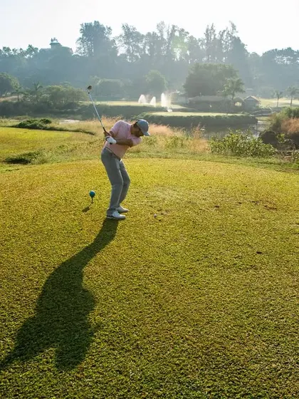 A player takes their shot from an elevated tee box, providing a great vantage point of the fairway below. The long shadow indicates a beautiful sunny day, perfect for a round of golf.