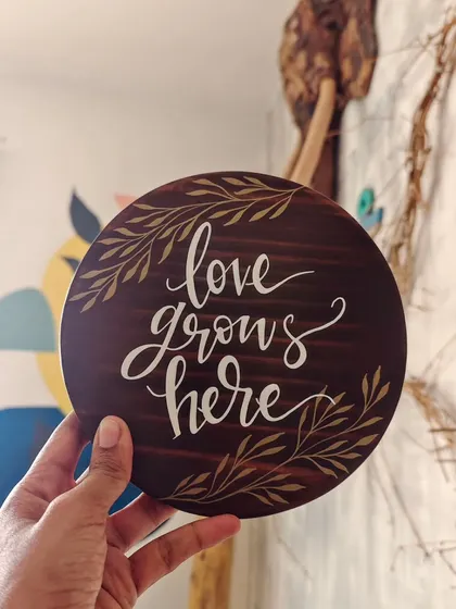 "Love grows here." This simple, elegant message is hand-painted on a round, dark-stained wooden plaque, perfect for celebrating a new home, a wedding, or the love in your family.