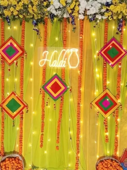 A close-up of a Haldi backdrop with a neon sign, showing the details of the hanging marigold garlands and colorful kite props against a lime green drape.