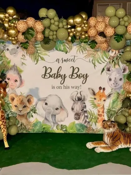 A gorgeous jungle-themed baby shower for a baby boy. The setup features a backdrop with watercolor animals, a balloon frame in sage green and gold, and large stuffed animals like a giraffe and tiger.