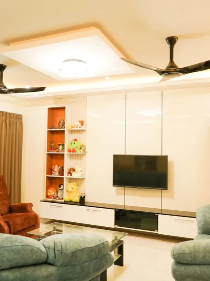 This bright and airy living room features a large, glossy white TV unit with an integrated display shelf. The multi-level false ceiling adds architectural interest and enhances the sense of space.