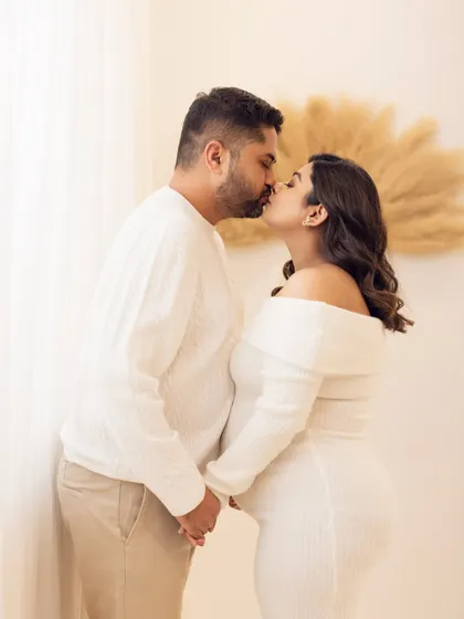 A passionate and romantic moment. This couple shares a kiss, holding hands in front of a softly lit window, their all-white outfits adding to the pure and tender feel of the photograph.