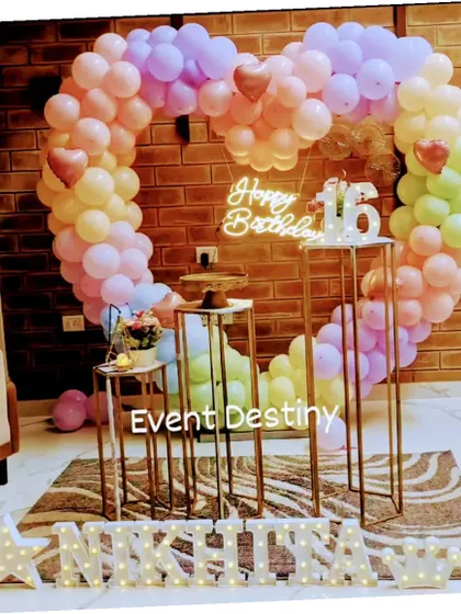A sweet sixteen birthday setup in a client's home, featuring a large heart-shaped balloon arch in pastel rainbow colors, a custom neon sign, and marquee letters for the name.