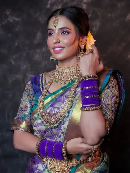 A smiling portrait of a model in a purple and green silk saree. Her pose, adjusting her bangles, feels natural and elegant, showcasing her traditional jewelry.