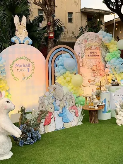 A whimsical Peter Rabbit-themed first birthday party. The main backdrop features storybook illustrations, pastel balloon garlands, and adorable rabbit props.
