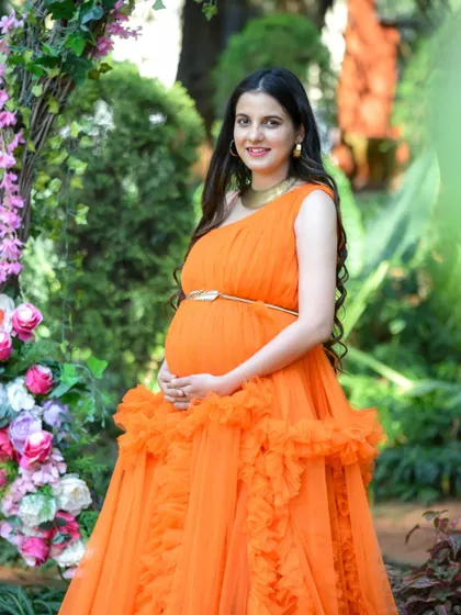 A classic portrait of a mom-to-be in a bright orange gown, standing in a beautiful garden setting.