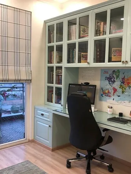 A bright and functional home office setup with a view. We designed this custom-built study unit in a light blue finish, featuring a spacious desk, drawers, and glass-fronted overhead cabinets for books and files.