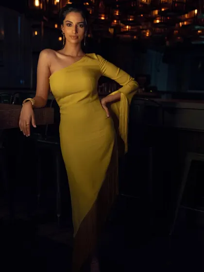 A full-length shot of a stunning yellow one-shoulder dress with fringe details. The styling is modern and sophisticated, set against a moody, ambient background.