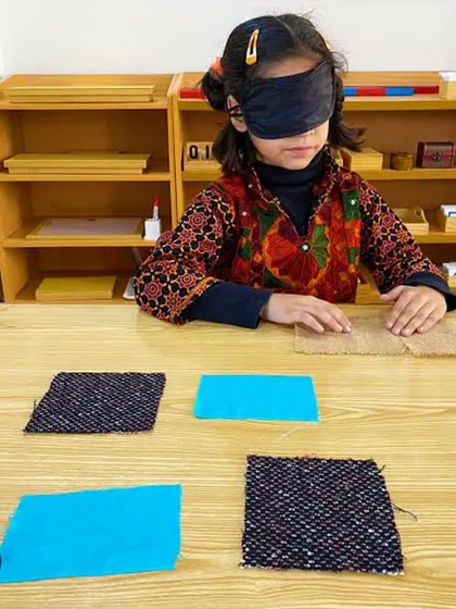 A blindfolded child uses her sense of touch to match different fabric textures. This classic Montessori sensorial activity refines the tactile sense, builds vocabulary for description, and develops concentration.