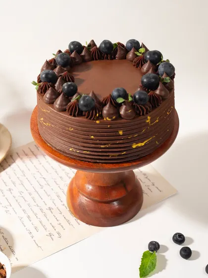 A full view of the Chocolate and Berry Cake on its wooden stand. The rich chocolate frosting and fresh blueberries make it a timeless classic.