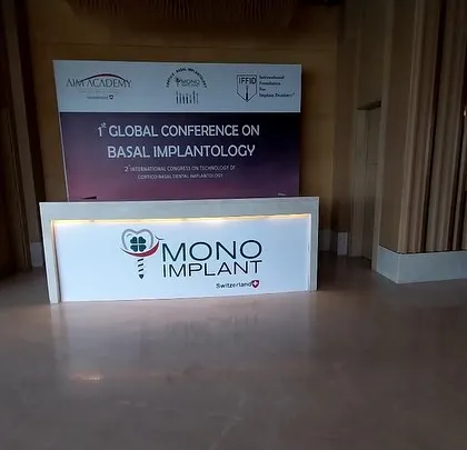 The main welcome counter at the Mono Implant trade show booth, positioned to greet attendees. The design is clean, well-lit, and prominently features the company logo for immediate brand recognition.