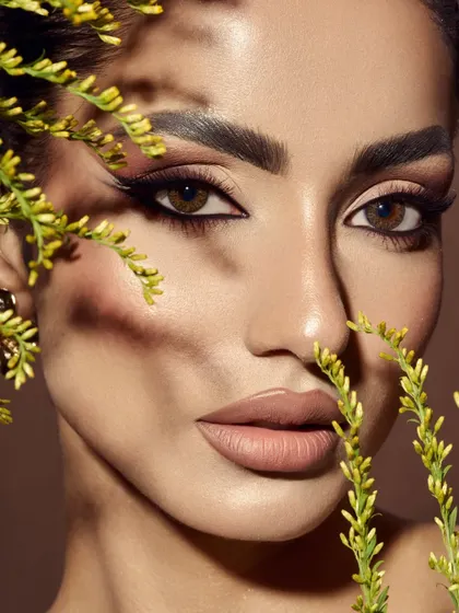 An artistic portrait using shadows and natural elements. The plant casts a pattern across the model's face, interacting with the sculpted makeup and creating a sense of mystery and depth.