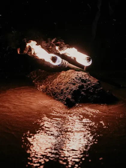 An atmospheric shot focusing on two flaming torches resting on a rock in the water. The reflection of the fire on the water's surface creates a warm, mesmerizing effect, perfect for setting a romantic scene.