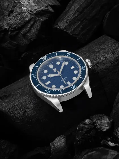A macro shot of a blue-faced watch on a bed of black charcoal. The contrast in texture and color makes the watch's details pop, giving it a rugged yet sophisticated look.
