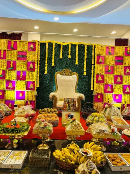 A full view of a Seemantham stage, featuring a colorful backdrop with hanging marigold garlands and brass bells. The foreground is filled with a wide variety of traditional sweets and offerings.