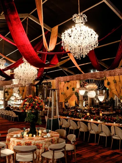 The dining area, with crystal chandeliers hanging beneath a canopy of draped red and gold velvet. The wooden floors and floral-print tablecloths create a rustic yet luxurious feel.
