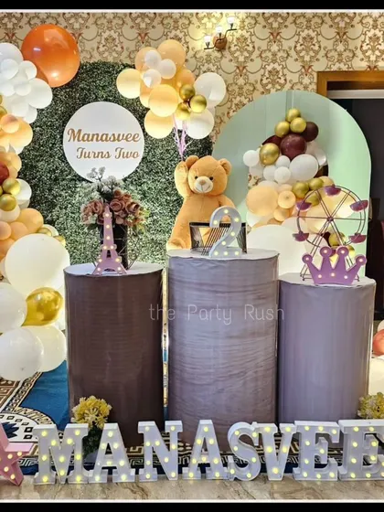 Manasvee's second birthday was a sweet teddy bear-themed party. The indoor setup features a teddy bear centerpiece, a Ferris wheel prop, and an earthy-toned balloon arch against a green hedge backdrop.