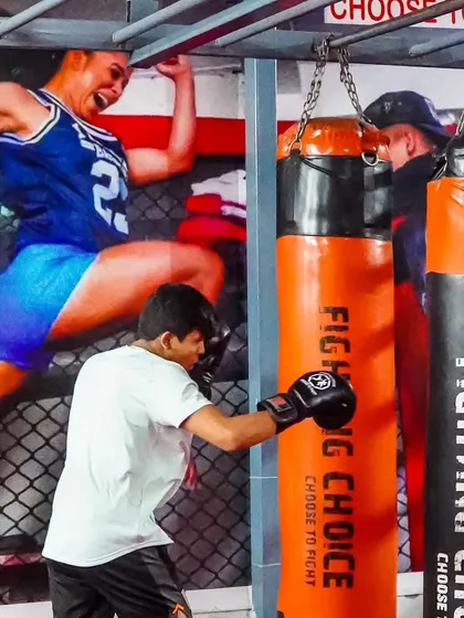 A young fighter practices his punches on the heavy bag, with an inspiring mural of a female warrior in the background. We train champions of all ages.