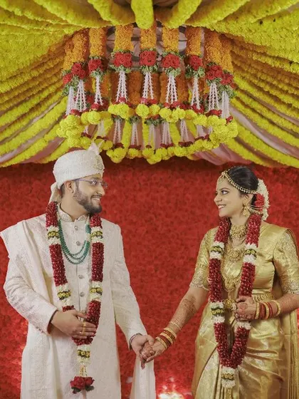 The couple stands before a grand, traditional backdrop of yellow marigolds and temple elephants, a perfect representation of a vibrant South Indian wedding.