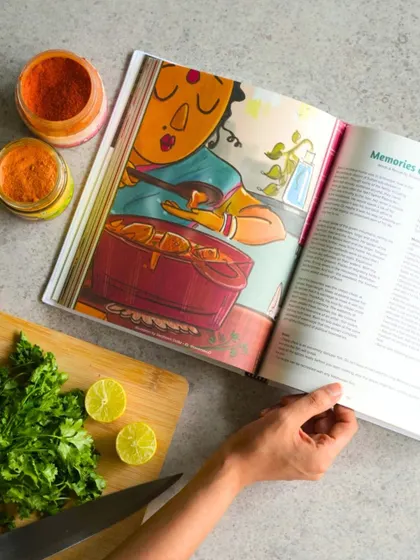 A hand holding our book 'Memories on a Plate', open to a vibrant illustration. This project transforms personal food remembrances into a beautiful culinary anthology.