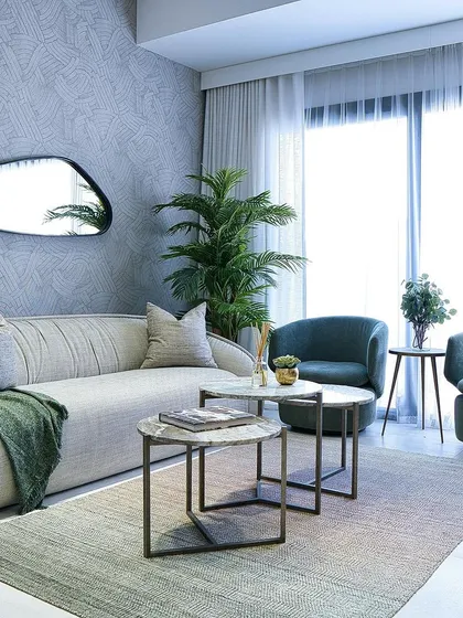 This living room features a unique, plush sofa with soft, rounded edges, paired with nested marble-top coffee tables and deep teal accent chairs, creating a comfortable yet stylish lounge area.