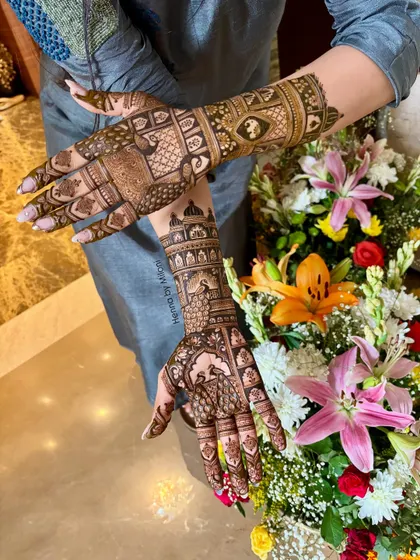 A beautiful shot of a bride's hands, adorned with detailed mehendi featuring peacocks and architectural elements, set against a bouquet of fresh flowers.