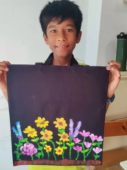 A young boy proudly holds up his tote bag, painted with a cheerful garden of yellow and purple flowers. My workshops encourage all children to explore their artistic side.