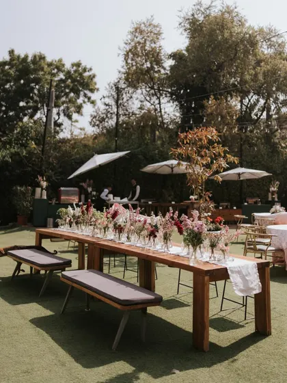 A long, rustic wooden table set for a daytime feast, adorned with simple wildflower arrangements in glass bottles.