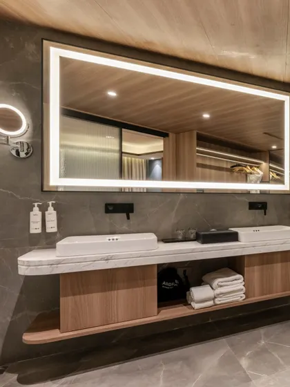 The dual-sink vanity in a guest suite bathroom, featuring a large, illuminated rectangular mirror. The combination of a floating marble countertop and light wood cabinetry creates a clean, spacious, and luxurious feel.