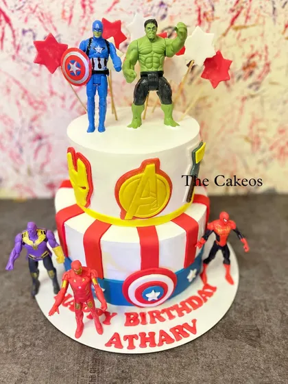 An action-packed two-tier Avengers theme cake for a superhero fan. The cake is decorated with logos of Iron Man and The Avengers, Captain America's shield, and topped with Hulk and Captain America figures.