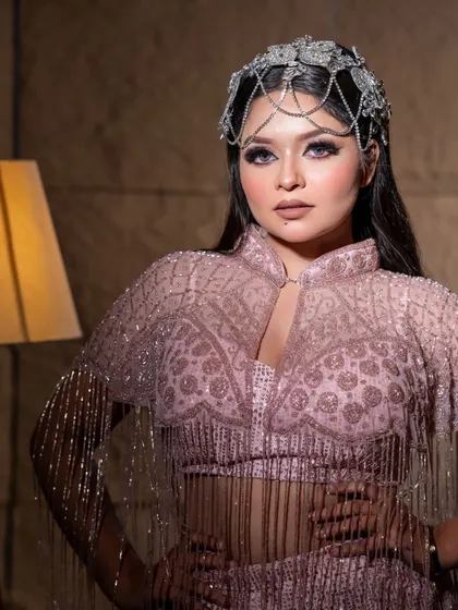 A model posing in a beautiful onion-pink gown with fringe details on the sleeves and a unique headpiece.