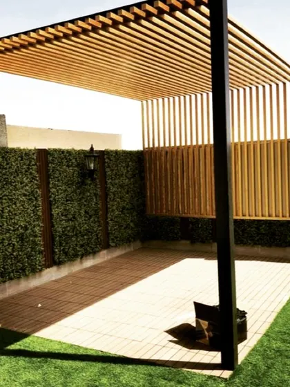 A modern pergola design combining a metal frame with a wooden slatted roof and side screen for partial privacy. The structure is set on a tiled patio next to an artificial turf lawn and a green wall.