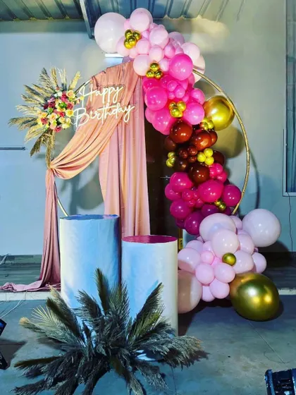A stylish birthday backdrop featuring a gold ring frame draped with pink fabric. The decoration is accented with a vibrant balloon garland in shades of pink and gold, and a "Happy Birthday" neon sign.