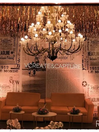A retro-style photo booth with a vintage newspaper print backdrop, orange sofas, and elegant chandeliers, perfect for a themed party.