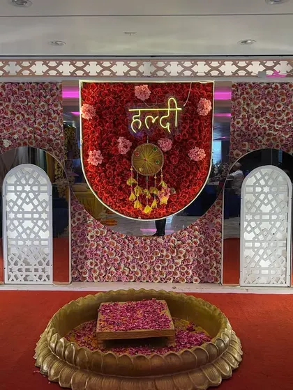 A grand and royal Haldi setup featuring a backdrop made entirely of red, pink, and yellow flowers, with decorative arches and a central lotus bowl.