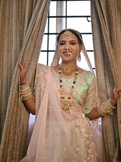 A playful peek from behind the curtains. Her makeup is fresh and bright, with a lovely pink lip that matches the dupatta.