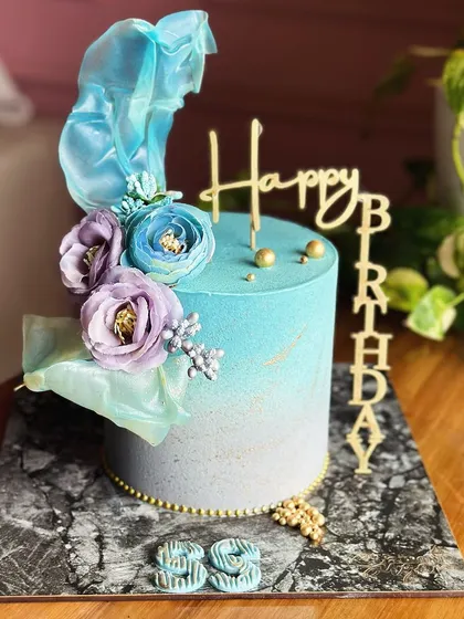 Elegance in every bite. This blue and lilac ombre cake is a dreamy blend of delicate hues, topped with sugar flowers and an ethereal sail for a magical birthday celebration.
